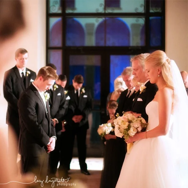 Bride, groom, and wedding guests bowing heads in prayer during a formal indoor ceremony with warm lighting.
